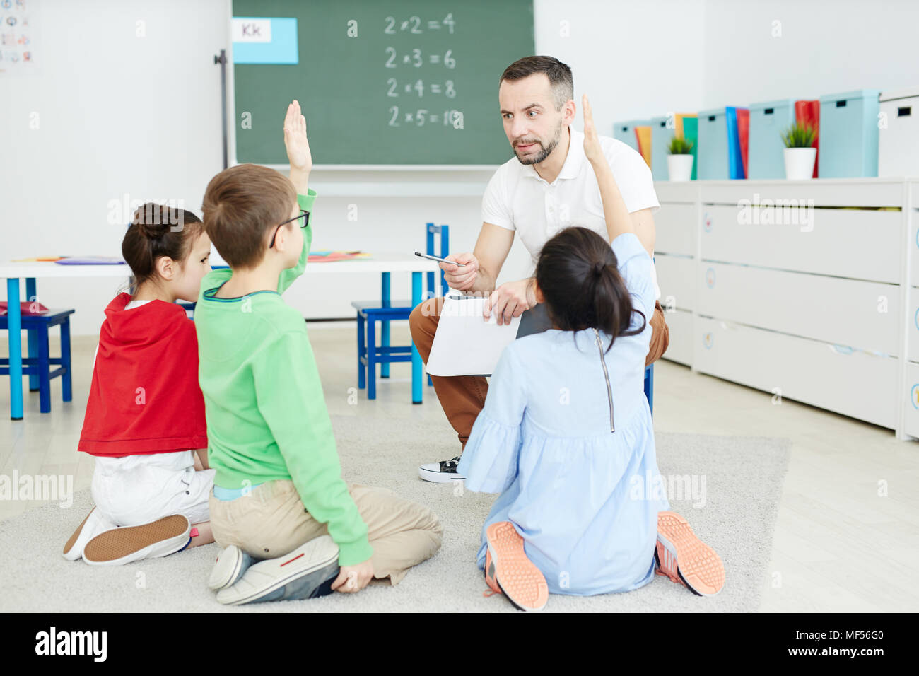 Studenti della scuola primaria alzando le mani e rispondere alle domande mentre è seduto sul pavimento e avente attività di matematica con insegnante maschio Foto Stock