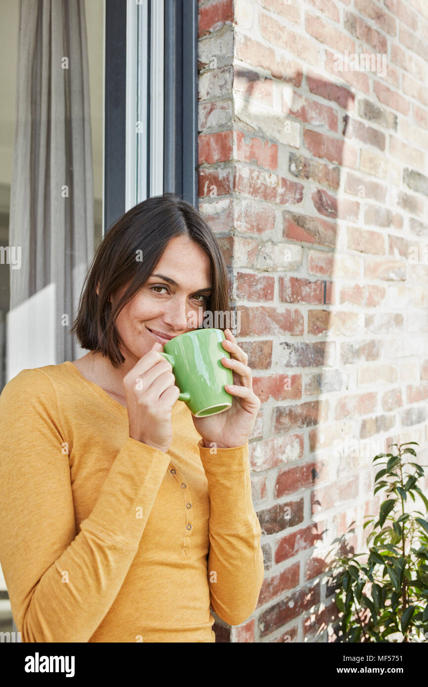 Ritratto di donna sorridente di bere dalla tazza di fronte la sua casa Foto Stock