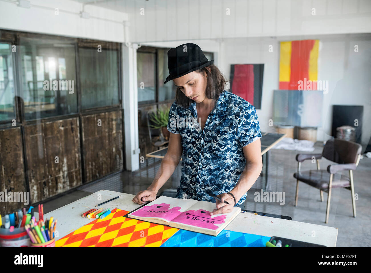 Artista al lavoro, disegno in un notebook nel suo loft studio con dipinti e opere d'arte in background Foto Stock