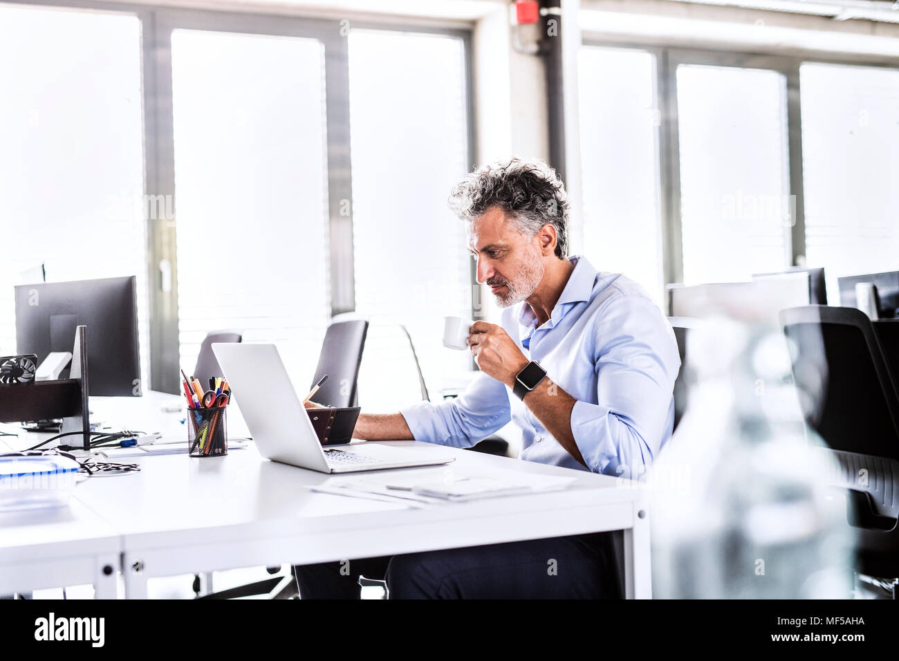 Imprenditore maturo seduto alla scrivania in ufficio utilizzando laptop e di bere il caffè Foto Stock