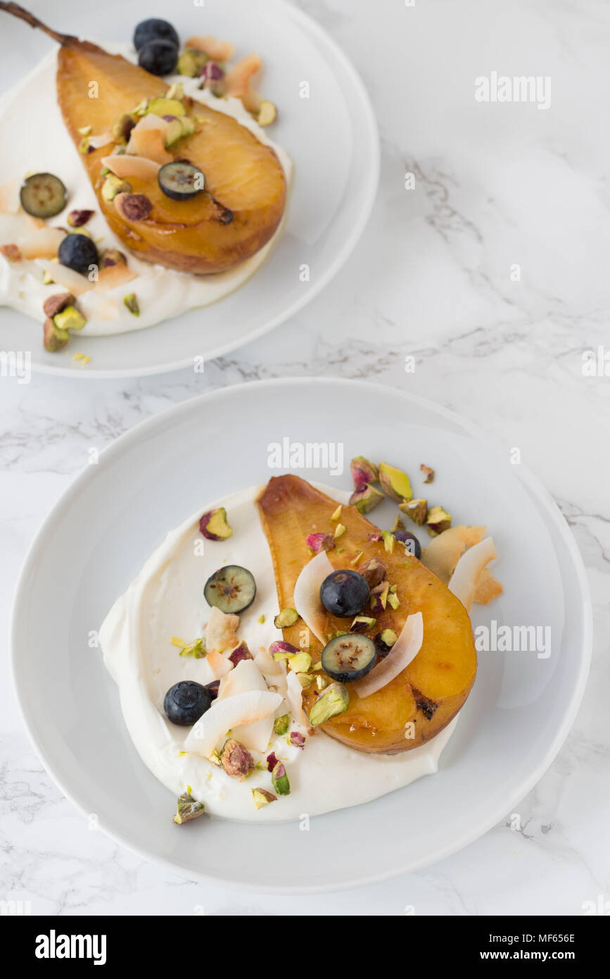 Due pere al forno con salsa di yogurt e decorate con pistacchi, scaglie di cocco e mirtilli. Le piastre bianche su sfondo marmo. Foto Stock