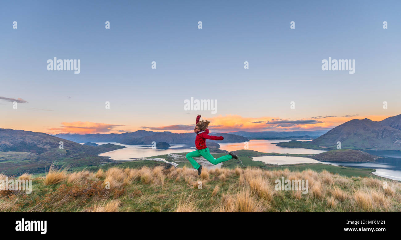Escursionista del salto in aria, la vista del Lago Wanaka e montagne, tramonto, picco roccioso, Glendhu Bay, Otago Southland, Nuova Zelanda Foto Stock