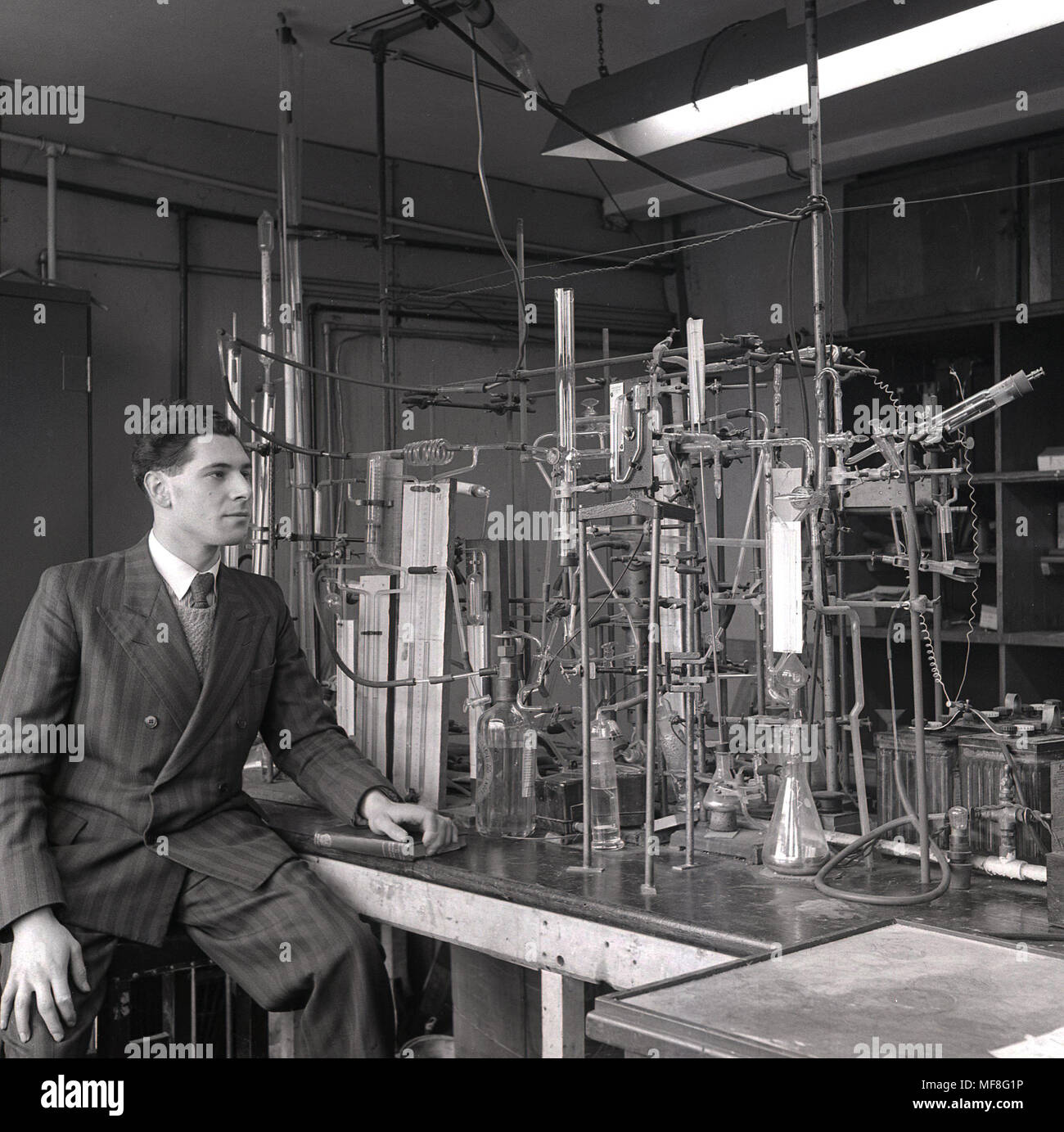 1948, storico, giovane maschio research scientist indossando un perno-striscia suit seduta accanto alla apparecchiatura di elaborati di tubazioni, tubi e bottiglie in vetro costruita per la sua ricerca sulla proprietà chimiche di carbone e di monossido di carbonio in un laboratorio nel dipartimento di carbone, gas e industrie di combustibile all Università di Leeds. Foto Stock