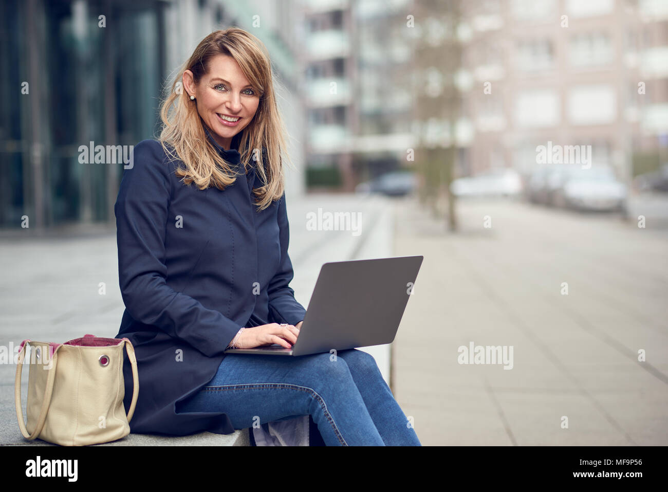 Attraente donna bionda usando il suo computer portatile in città seduti sui gradini davanti a un edificio commerciale sotto il sole sorride mentre lavora Foto Stock