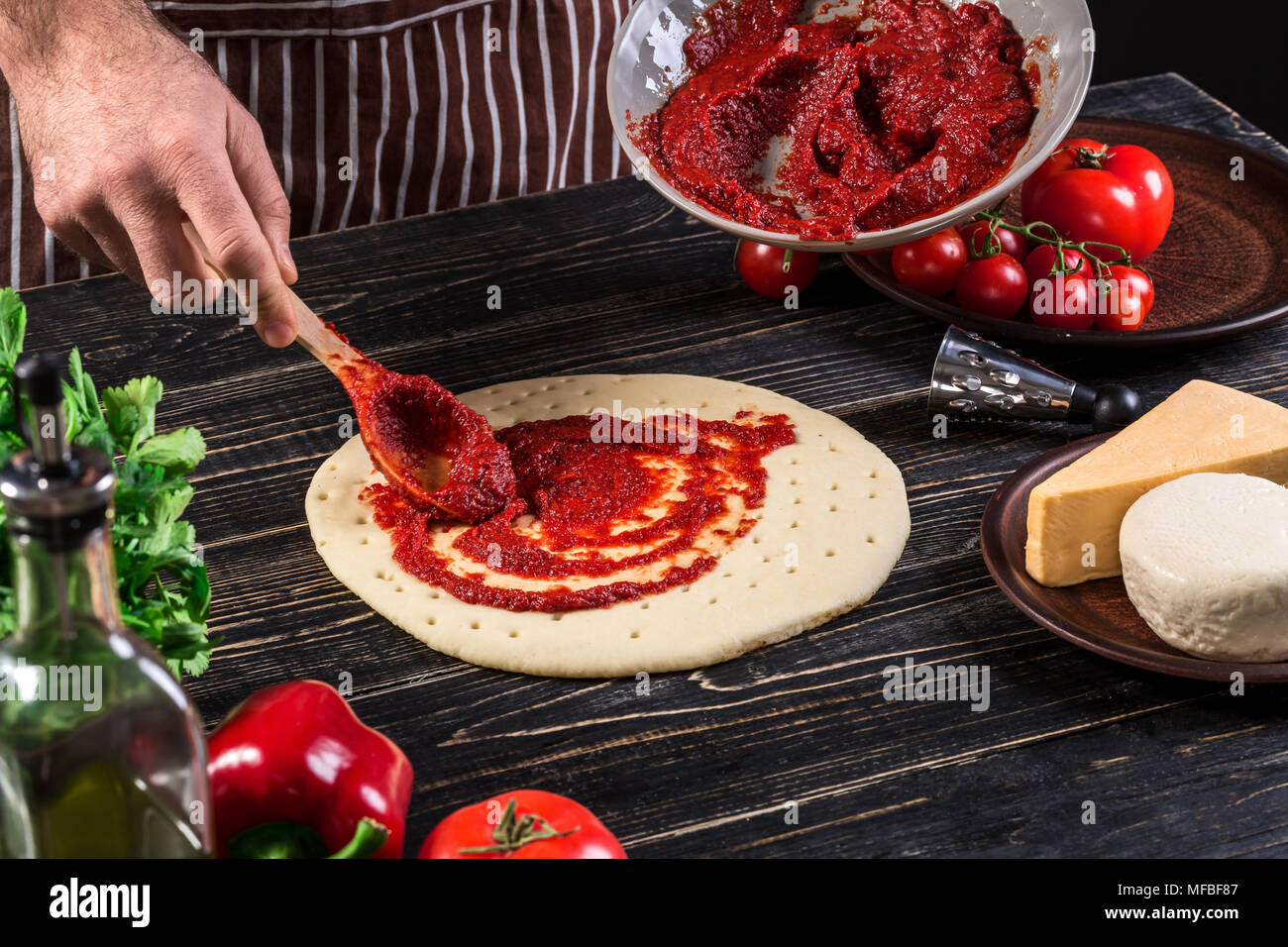 Un maschio di spargimento a mano la salsa di pomodoro sulla base per pizza con cucchiaio su un vecchio sfondo di legno Foto Stock