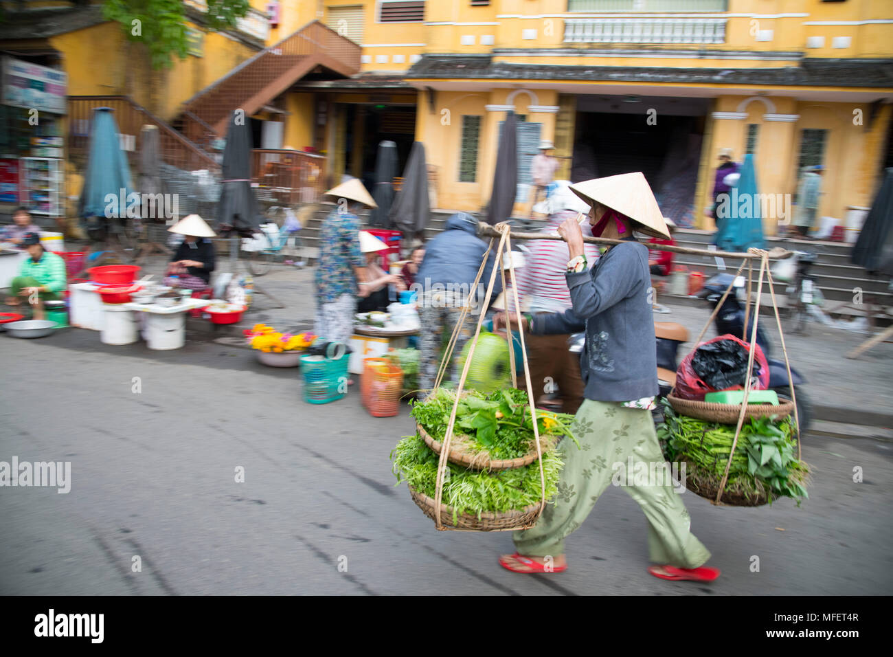 Una donna a piedi attraverso il mercato con pesanti ceste di ortaggi in Hoi An, Quang Nam Provincia, Vietnam Foto Stock