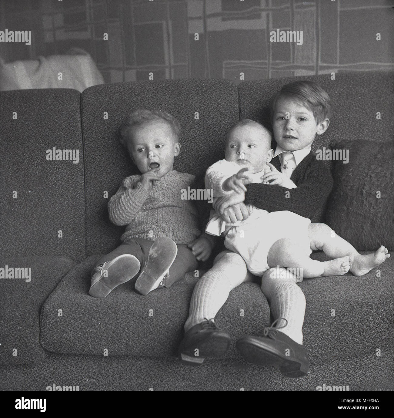 Degli anni Cinquanta, storico, tre fratelli seduti insieme su un divano, con uno dei ragazzi piccoli, il più antico, tenendo il suo fratellino nelle sue braccia, Inghilterra, Regno Unito. Foto Stock