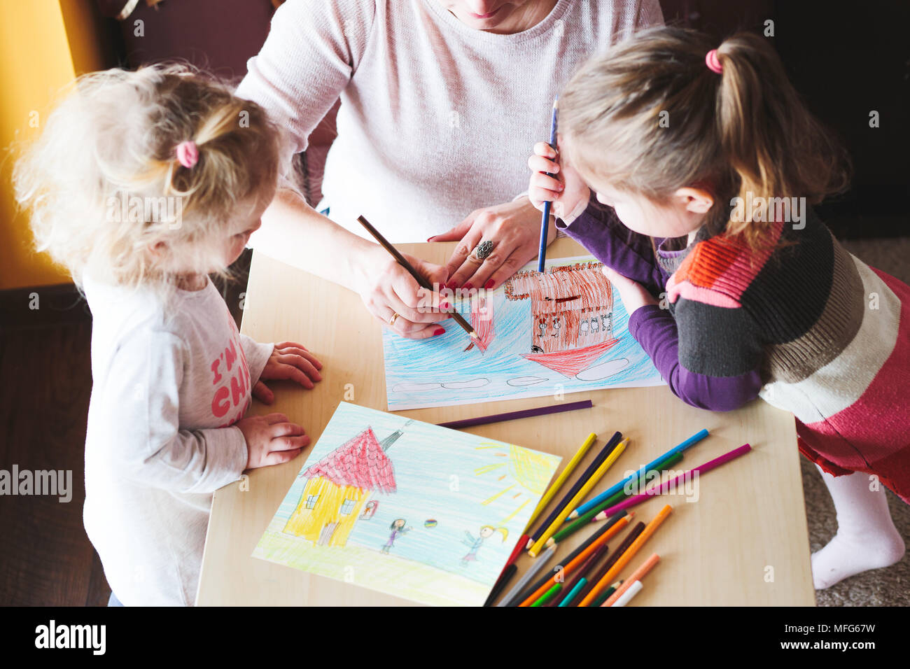 Mom con bambine il disegno di una foto a colori di casa e giocare con i bambini usando matite permanente al tavolo nella sala al coperto. Ripresa dall'alto Foto Stock