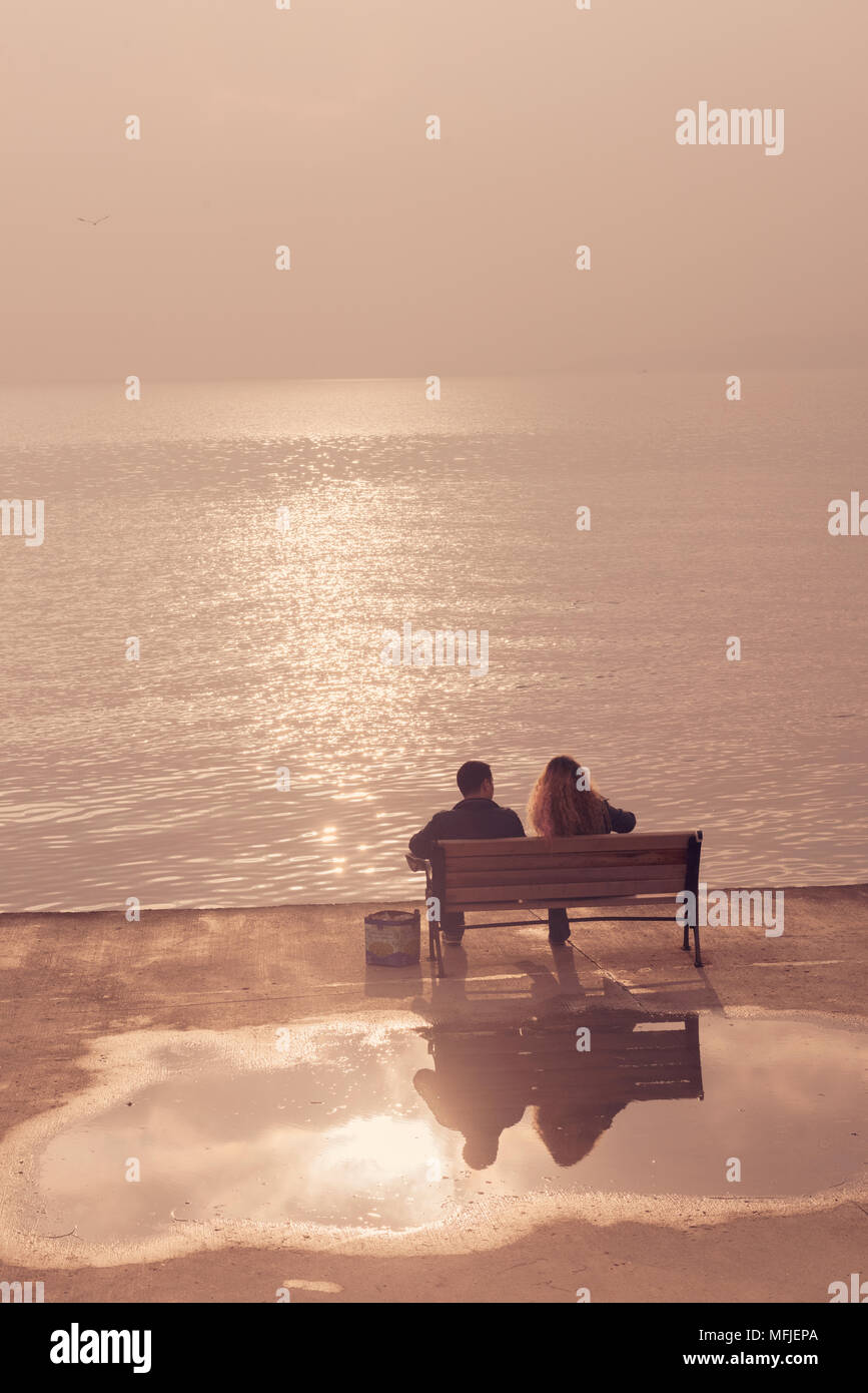 Una coppia seduta su una panchina vicino all'acqua, guardando il tranquillo riflesso del tramonto, con luce soffusa che crea un'atmosfera tranquilla e romantica. Foto Stock