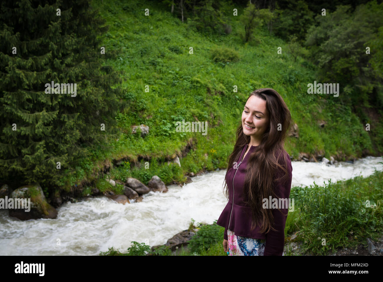 Giovane donna sorge nel parco nazionale sullo sfondo di una verde collina con il fiume di montagna nella giornata d'estate. Ragazza alla moda che guarda lontano e sorridente. Foto Stock