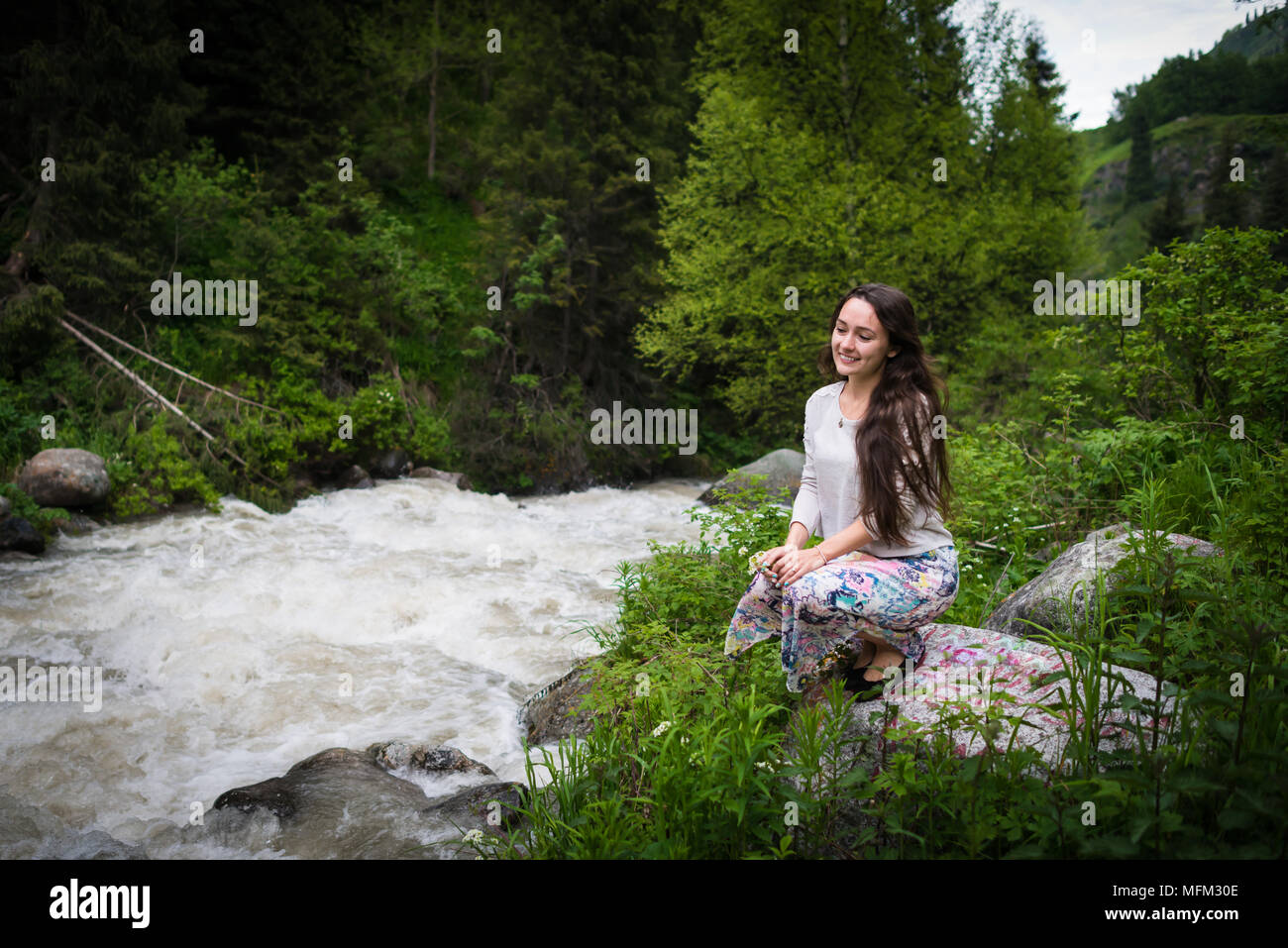 Giovane donna siede su pietra nel parco nazionale e si affaccia sulla montagna di ebollizione fiume. Ragazza che guarda lontano e sorridente su sfondo di colline verdi e alberi Foto Stock