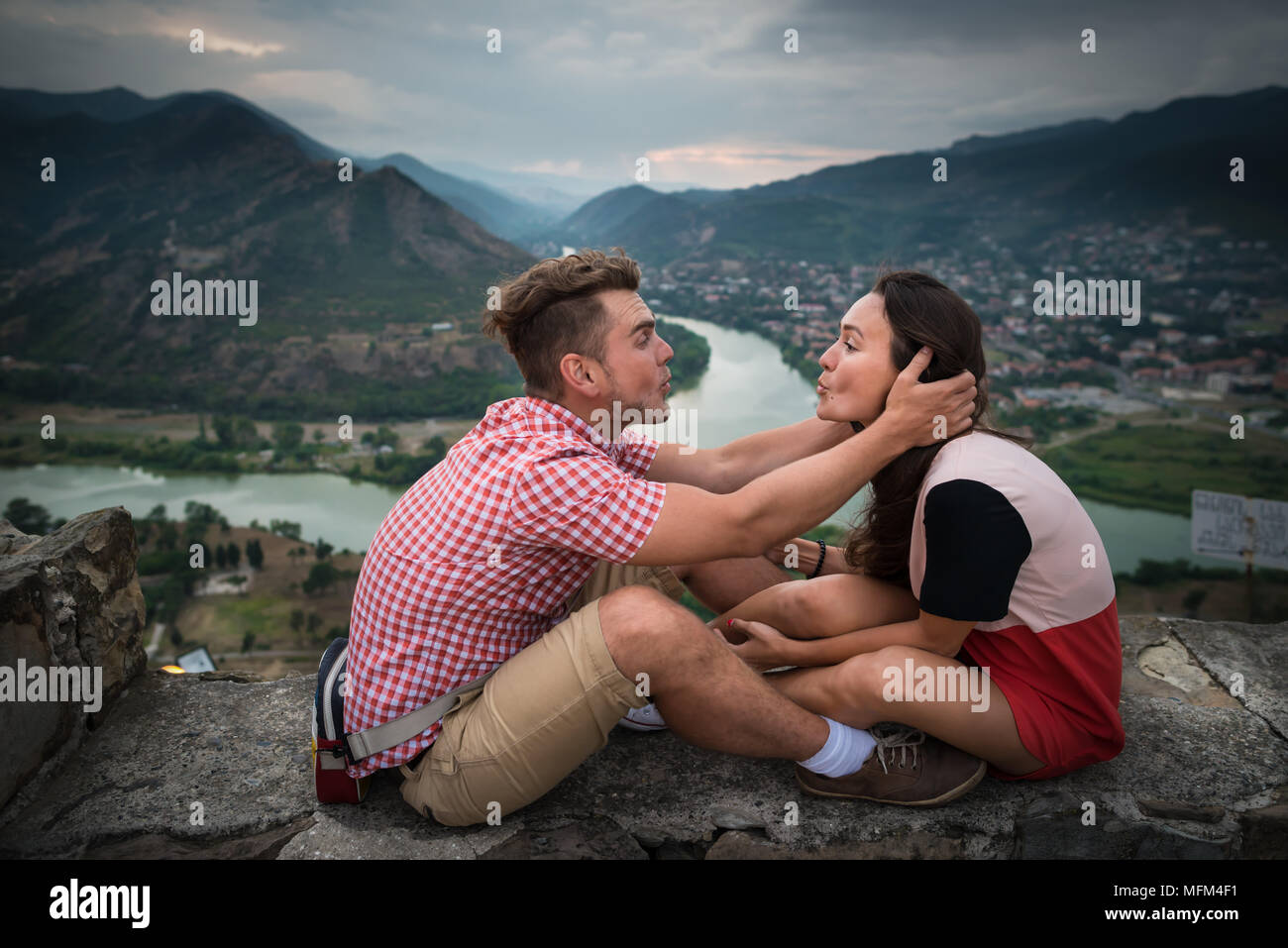 Matura in amore si siede sulla pietra sullo sfondo di montagne, il fiume e la città piccola al piano di sotto. Giovane uomo baci pretty woman. Foto Stock