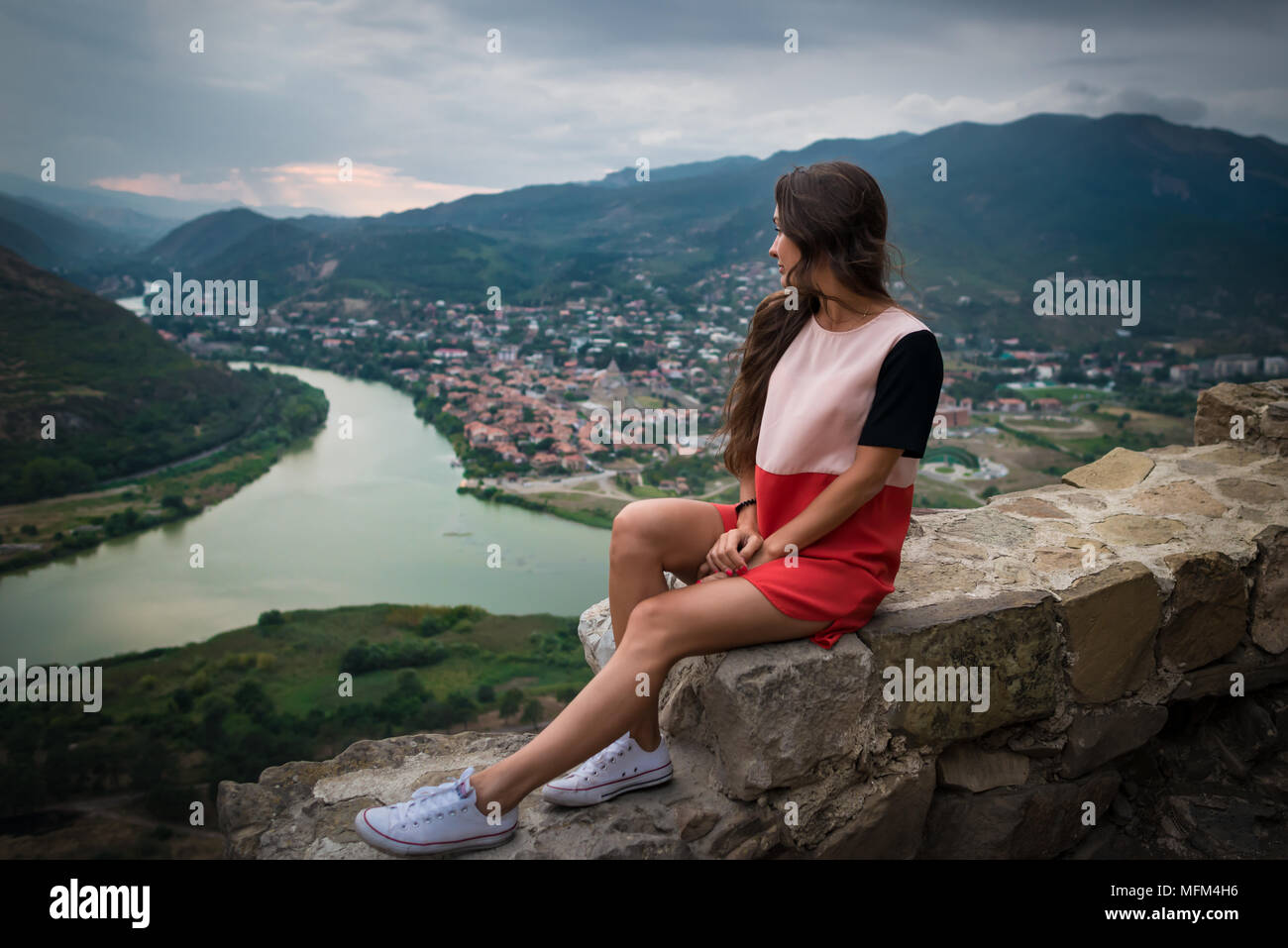 Giovane donna elegante siede sulla pietra sullo sfondo di montagne, il fiume e la città piccola al piano di sotto. Bella ragazza cerca in lato. Destinazione di viaggio. Foto Stock