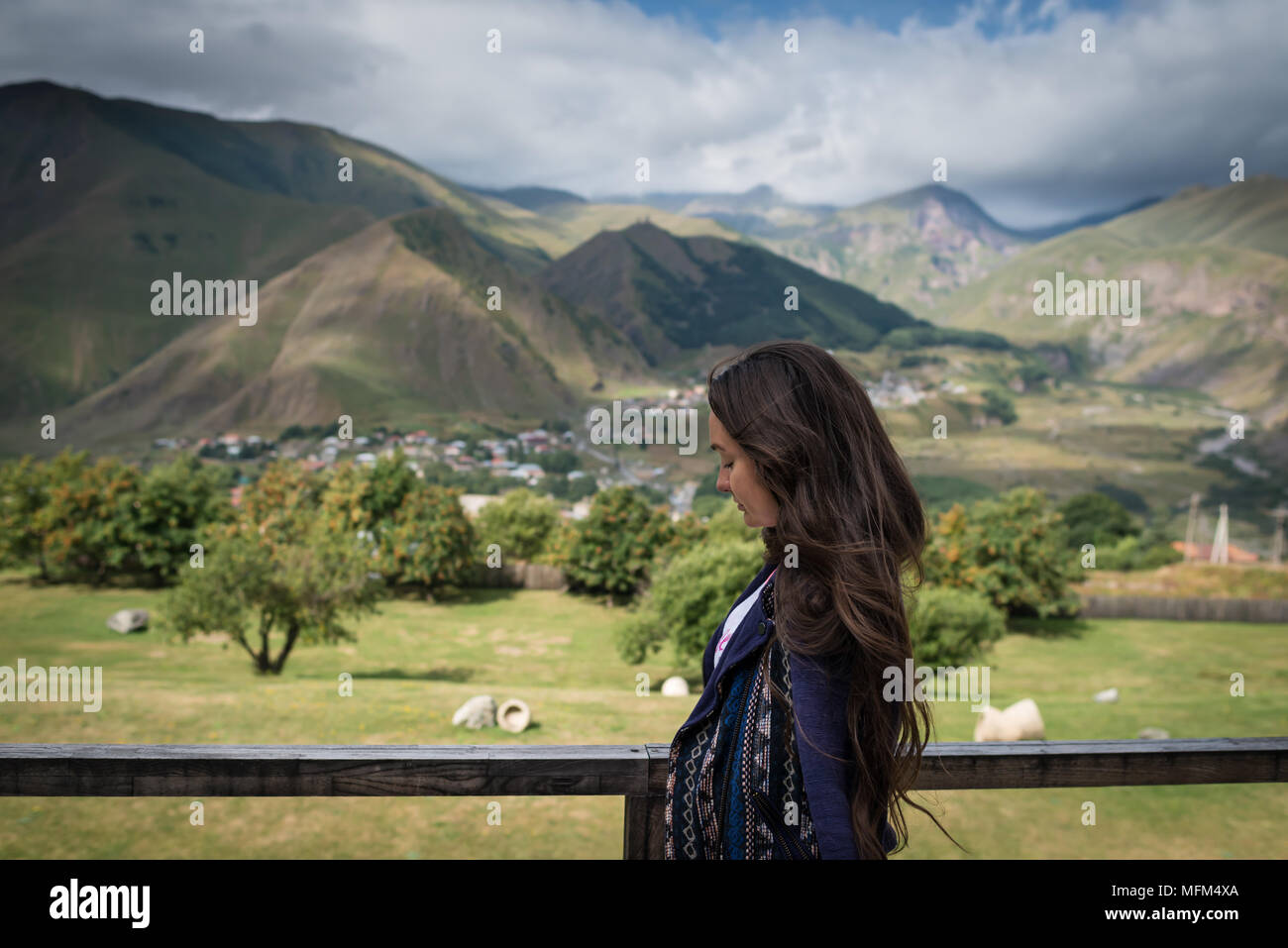 Giovane donna pensieroso con gli occhi chiusi si erge sullo sfondo di montagne e piccolo villaggio nella valle. Vista dal lato sulla ragazza. Estate giornata di sole. Foto Stock