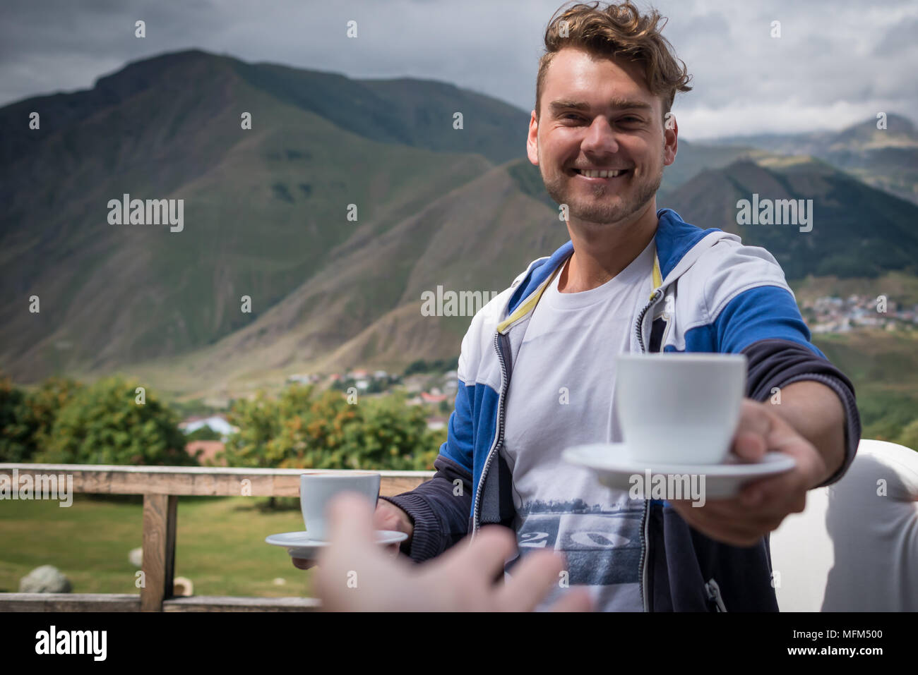 Giovane uomo tiene fuori una tazza di caffè. Felice ragazzo con bevande al mattino sulla terrazza con bella vista sulle montagne. Buona mattina concetto. Foto Stock