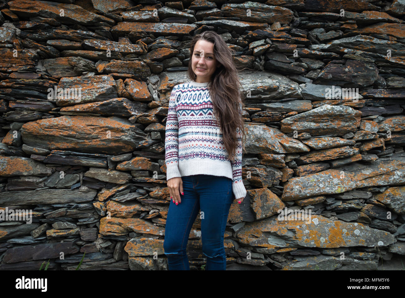 Bella donna sorge isolato su sfondo di pietre. Vista dal lato anteriore. Elegante ragazza in maglione e jeans sta cercando nella fotocamera Foto Stock