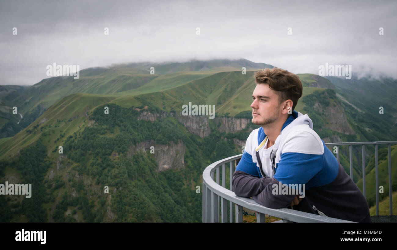 Giovane uomo sorge alla piattaforma di osservazione sullo sfondo delle bellissime montagne verdi e il cielo. Ragazzo bello avere un periodo di riposo prima di escursionismo. Foto Stock