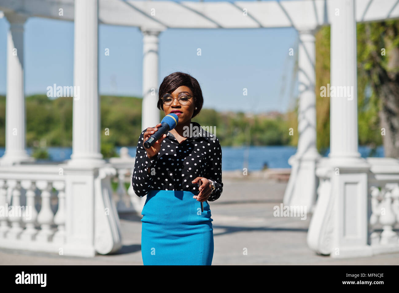 Elegante americano africano ragazza modello presentatore TV con microfono in bicchieri blu gonna e Blusa nera poste all aperto contro il bianco arco in pietra con m Foto stock Alamy