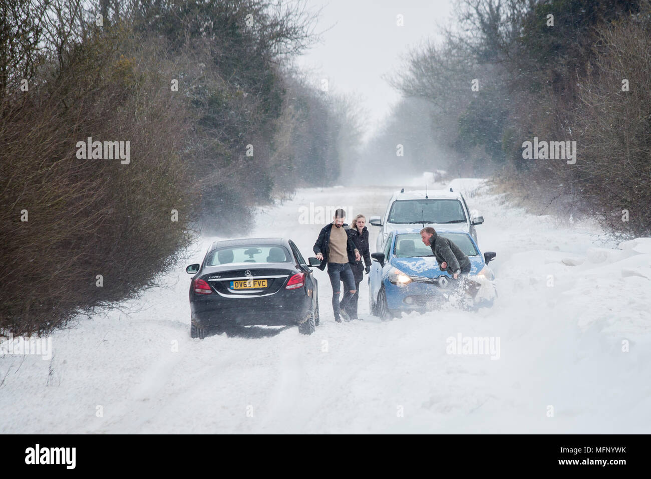 Un uomo con una vanga per scavare la sua vettura al di fuori di un snow drift in piedi nella neve profonda che egli ha per la battaglia è il forte vento con raffiche fino in tutto il paese strada come altri veicoli di passaggio. Foto Stock