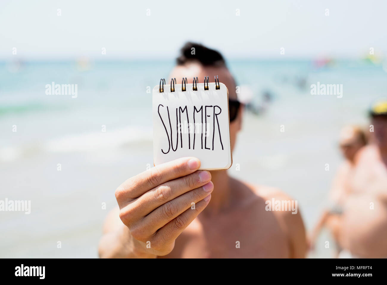 Primo piano di un giovane uomo caucasico in spiaggia che mostra una spirale notepad con la parola manoscritta estate in prima pagina Foto Stock