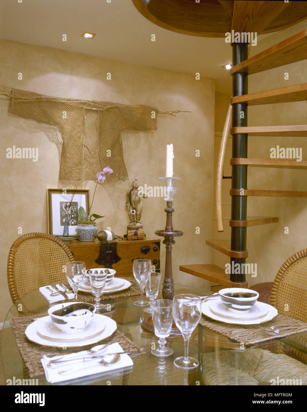 Paese sala da pranzo con trama pareti di gesso, floating scalinata a spirale e un top in vetro tavolo da pranzo con sedie di vimini. Foto Stock