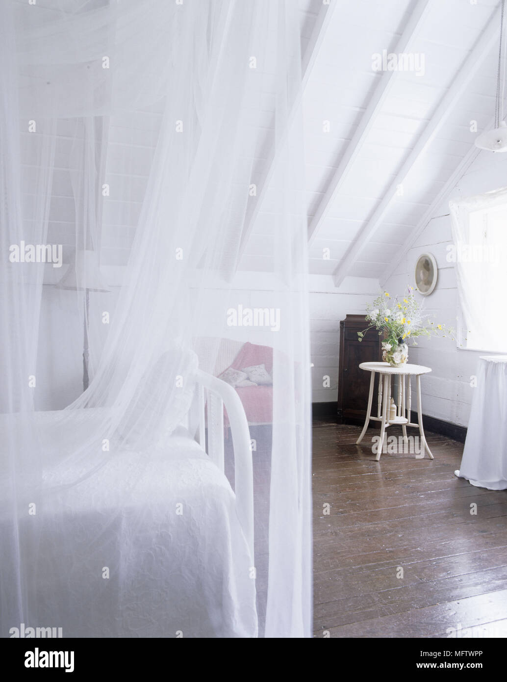 Dettagli di una tradizionale camera da letto con un bianco tetto spiovente e pavimenti in legno scuro con un letto ricoperto di netting e biancheria da letto bianca con un piccolo tabl Foto Stock
