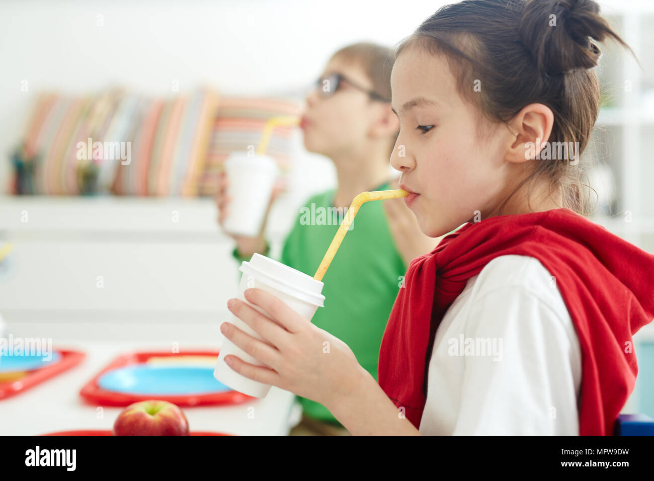 Bella ragazza caucasica seduta a tavola nella scuola primaria mensa e di bere succo di frutta dopo pranzo su sfondo sfocato, vista laterale Foto Stock