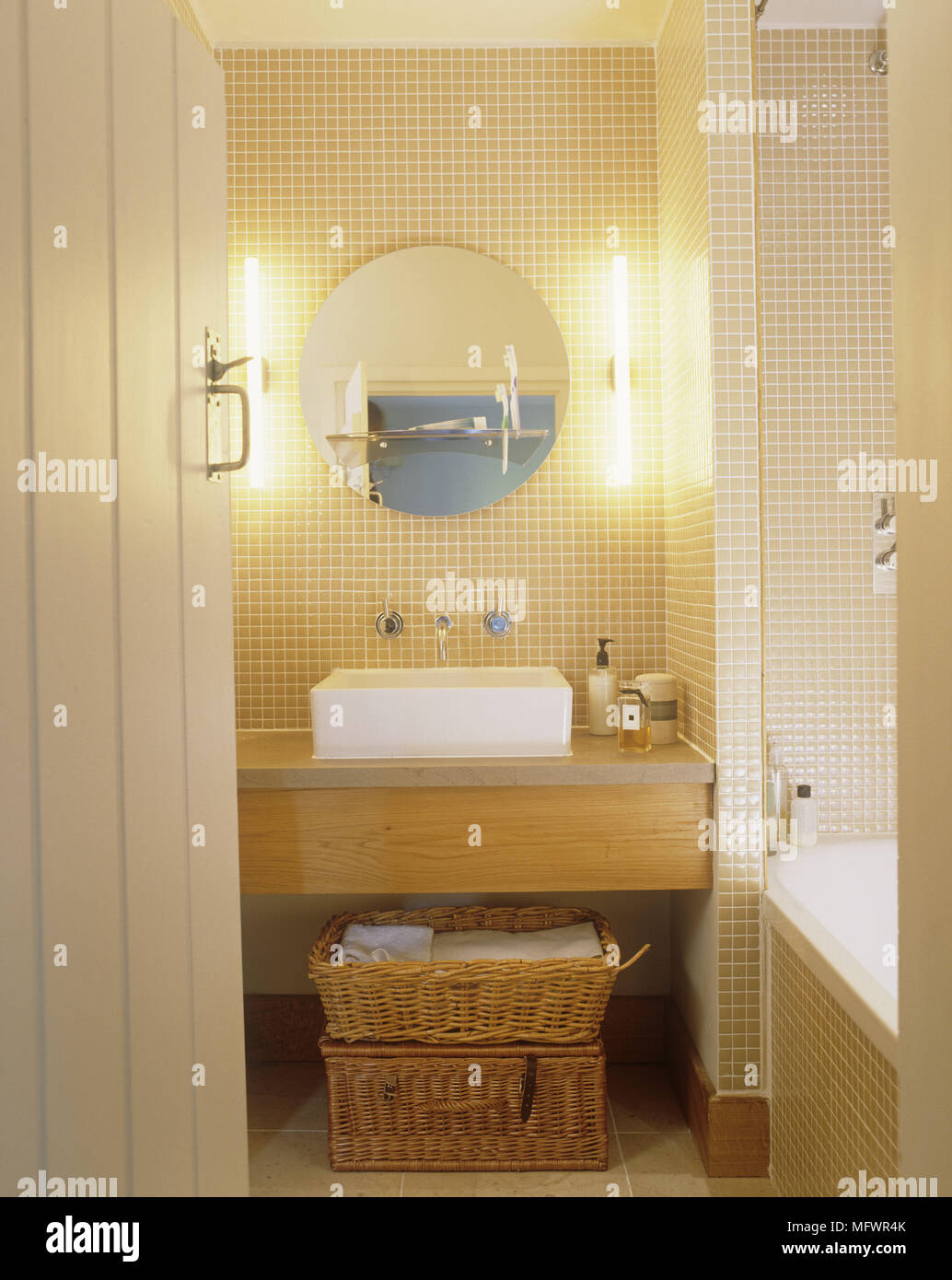 Bagno con pareti piastrellate e specchio circolare sopra il lavabo sulla scatola di legno con cestini di vimini al di sotto di Foto Stock
