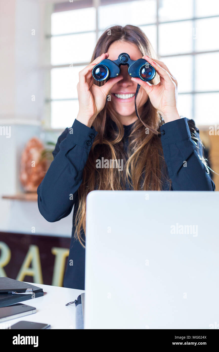 Deutschland, München, Ritratto Geschäftsfrau im Büro vor Laptop durch Fernglas schauend Foto Stock