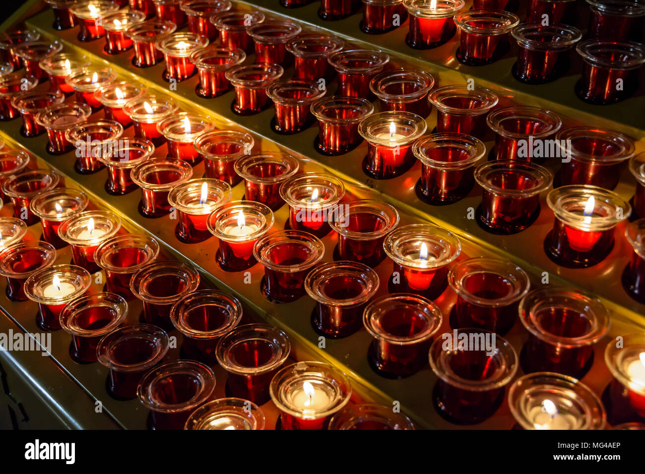 Bianco di piccole candele votive illuminato e posto in rosso candelieri di vetro fra quelle vuote, allineati su una candela titolare di stand in una chiesa cattolica. Foto Stock