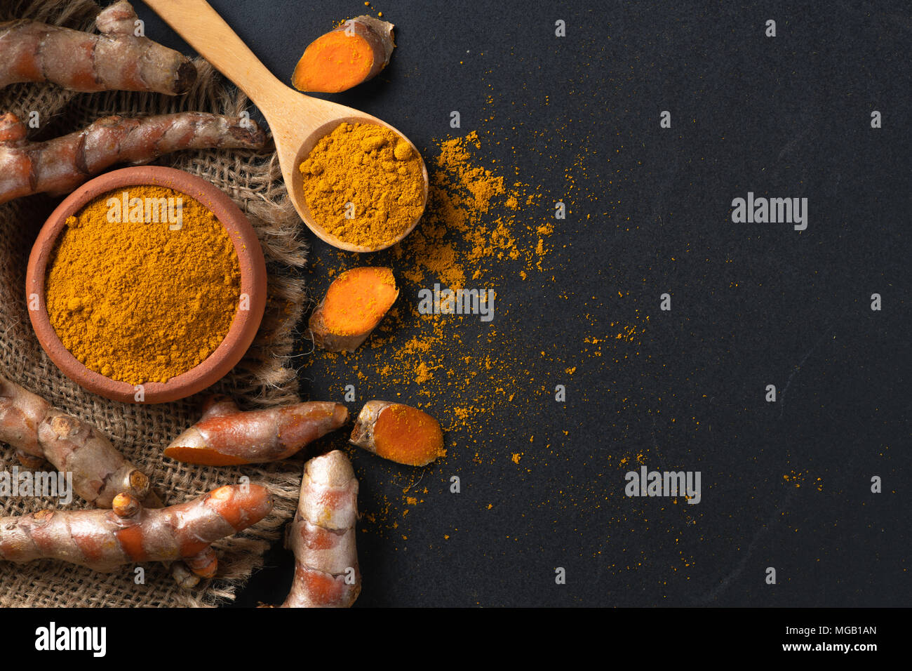 La curcuma in polvere in cucchiaio di legno con la curcuma root su tavola nera, le spezie e le erbe concept Foto Stock