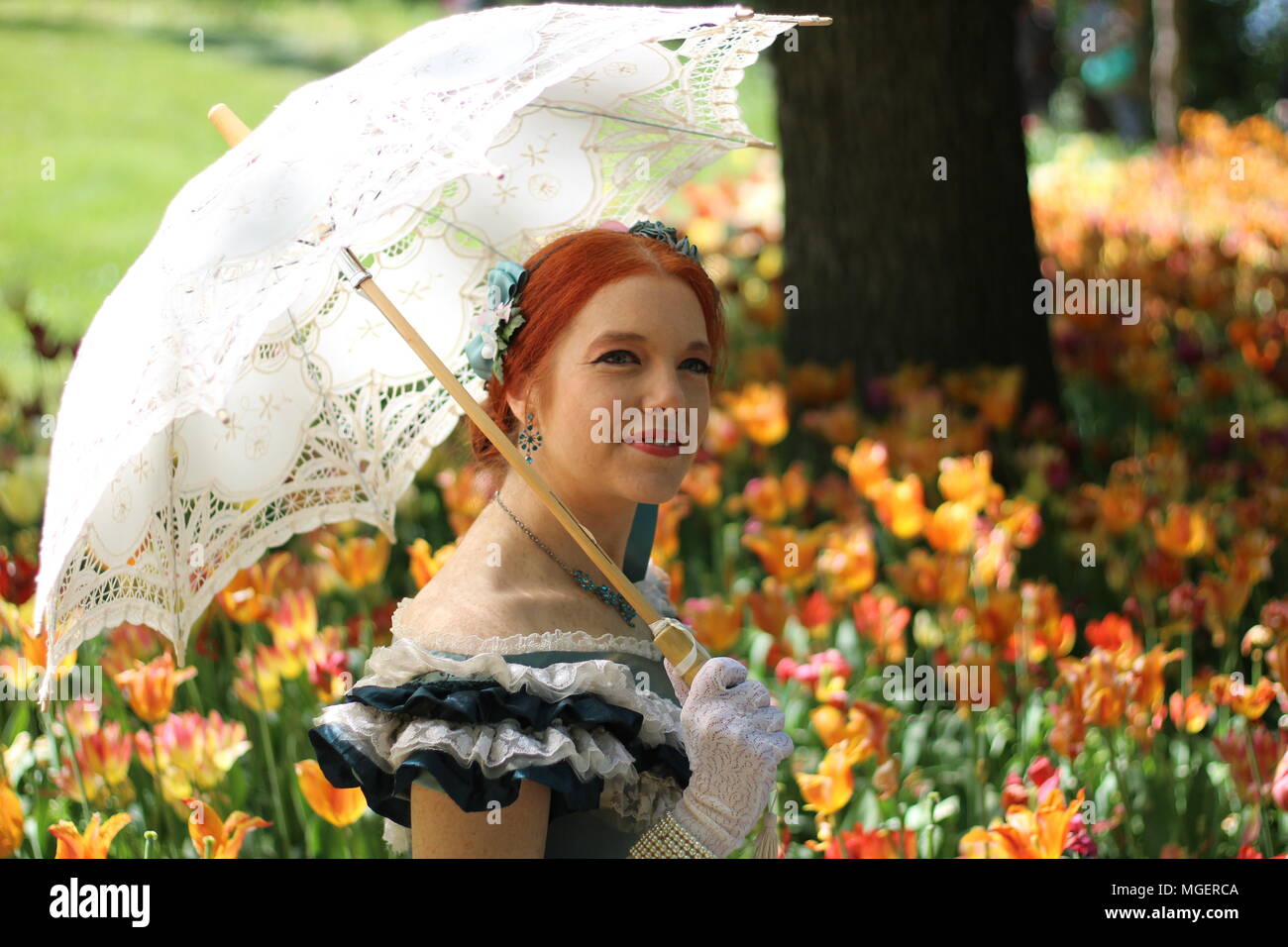 Una donna con i capelli rossi e un ombrellone bianco per proteggersi dal sole si rilassa in un campo di rosso e tulipani gialli Foto Stock