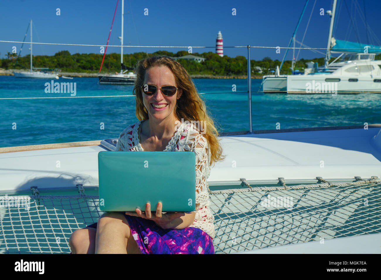 Giovane donna nel cappello per il sole e occhiali da sole funziona su un  computer portatile dalla prua di una barca a vela, barche e il faro in  background Foto stock -