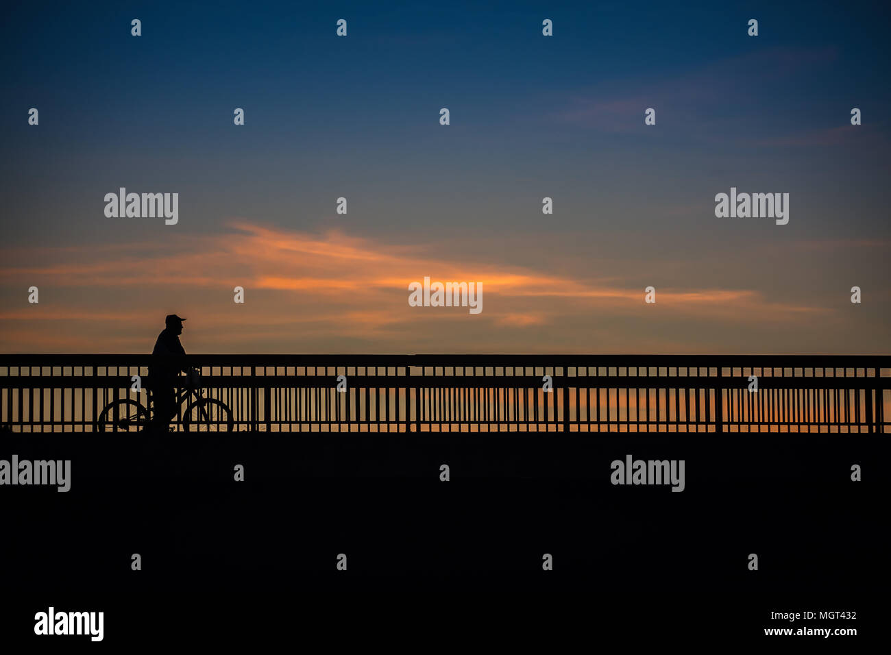 Gli uomini anziani è in sella ad una bicicletta su un ponte, con incredibile cielo blu e nuvole in background al tramonto, silhouette Foto Stock