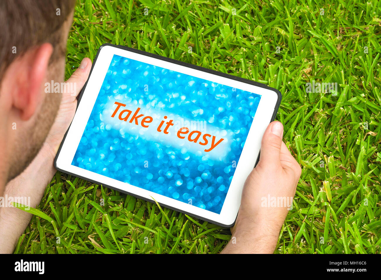 Giovane uomo disteso su erba in giardino con dispositivo tablet guardando sfocato sfondo blu con la frase "Take it easy' scritto su di esso. Foto Stock