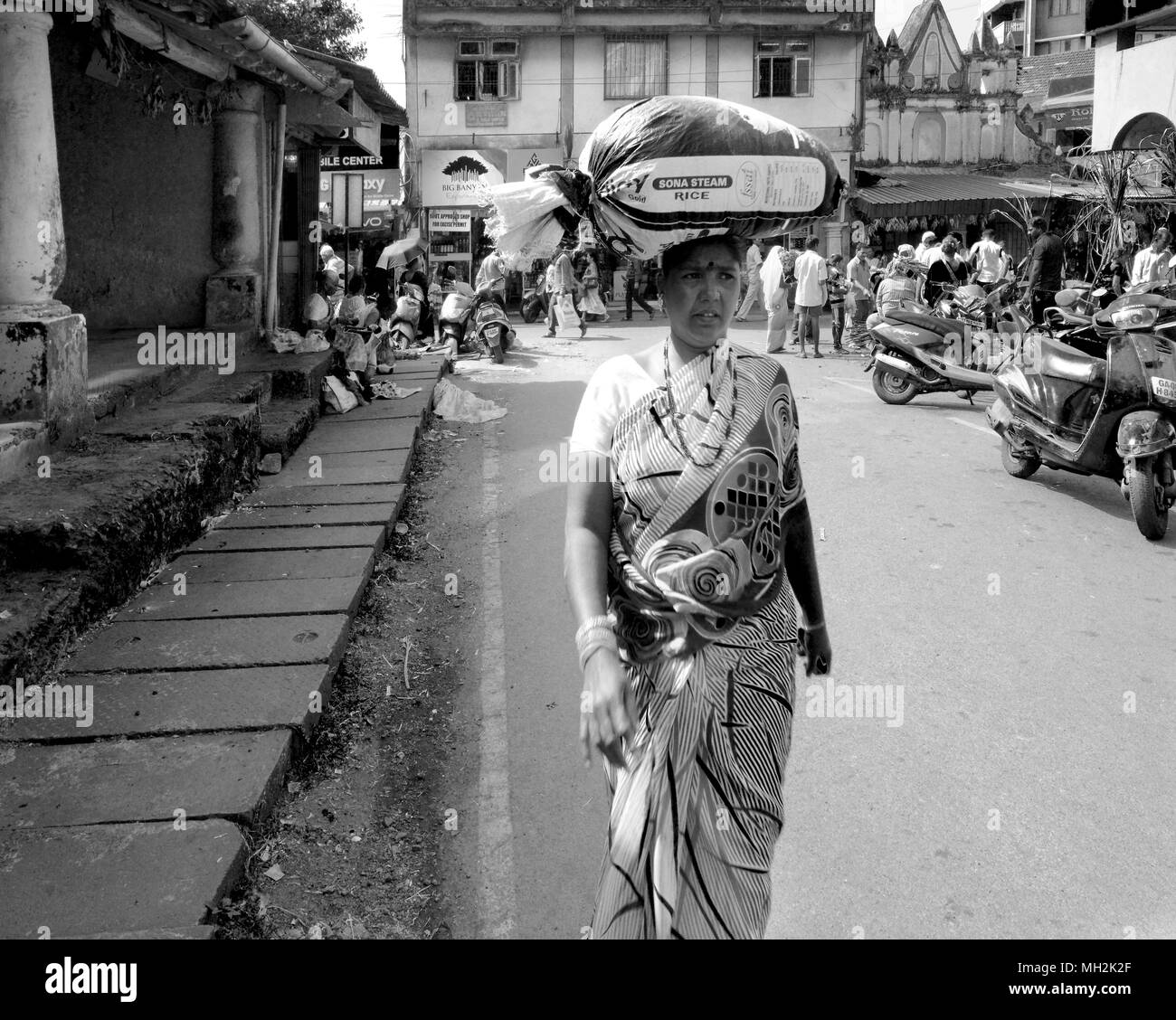 Una donna che porta una grande borsa pesante di riso bilanciato sulla sua testa attraverso la strada di Margao, vicino Benaulim, Goa, India Foto Stock