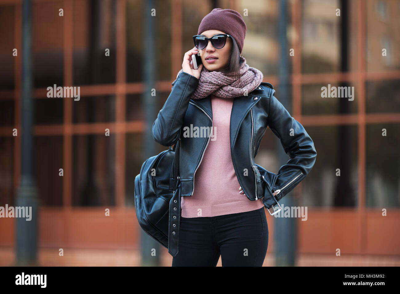 Moda giovane donna in nero giacca di pelle utilizzando il cellulare in una strada di città. Elegante modello femminile di indossare occhiali da sole beanie e sciarpa per esterno Foto Stock