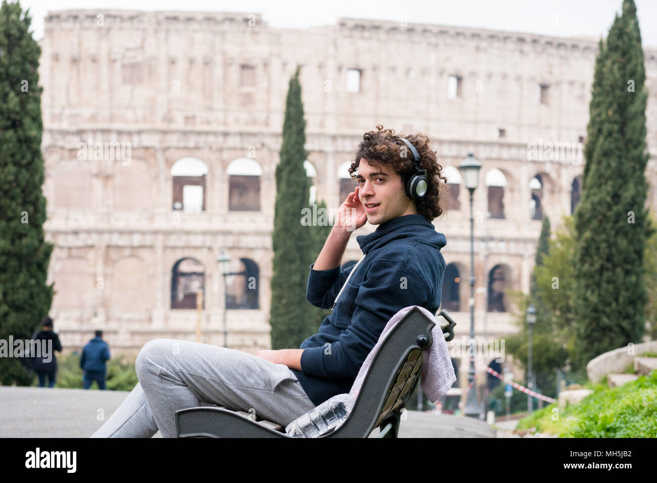 Bel giovane nei panni dello sport ascoltando musica seduto su una panchina con il Colosseo in background. Giovane atleta in tuta rilassante dopo Foto Stock