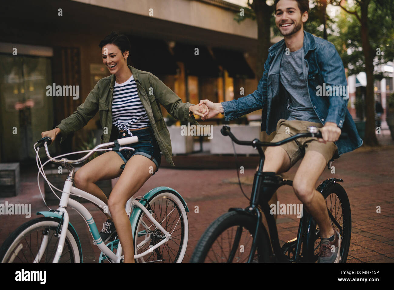 Felice coppia giovane Holding Hands e passeggiate in bici. Uomo sorridente e la donna in bicicletta assieme nella città. Foto Stock