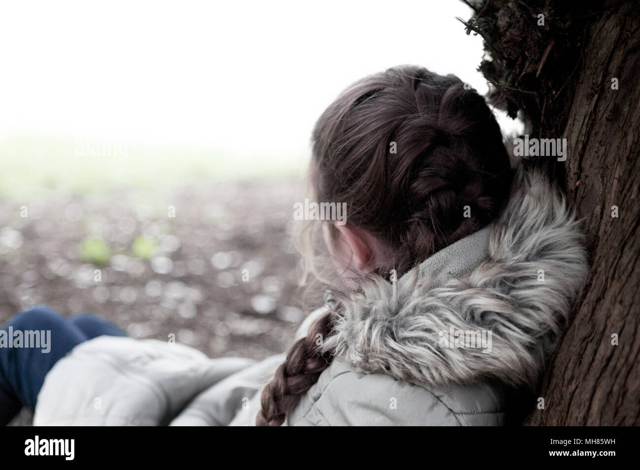 Una giovane ragazza (di età compresa tra otto), avvolto a caldo in una pelliccia cappotto, si appoggia contro un albero con la schiena per la telecamera in un crollati pongono Foto Stock