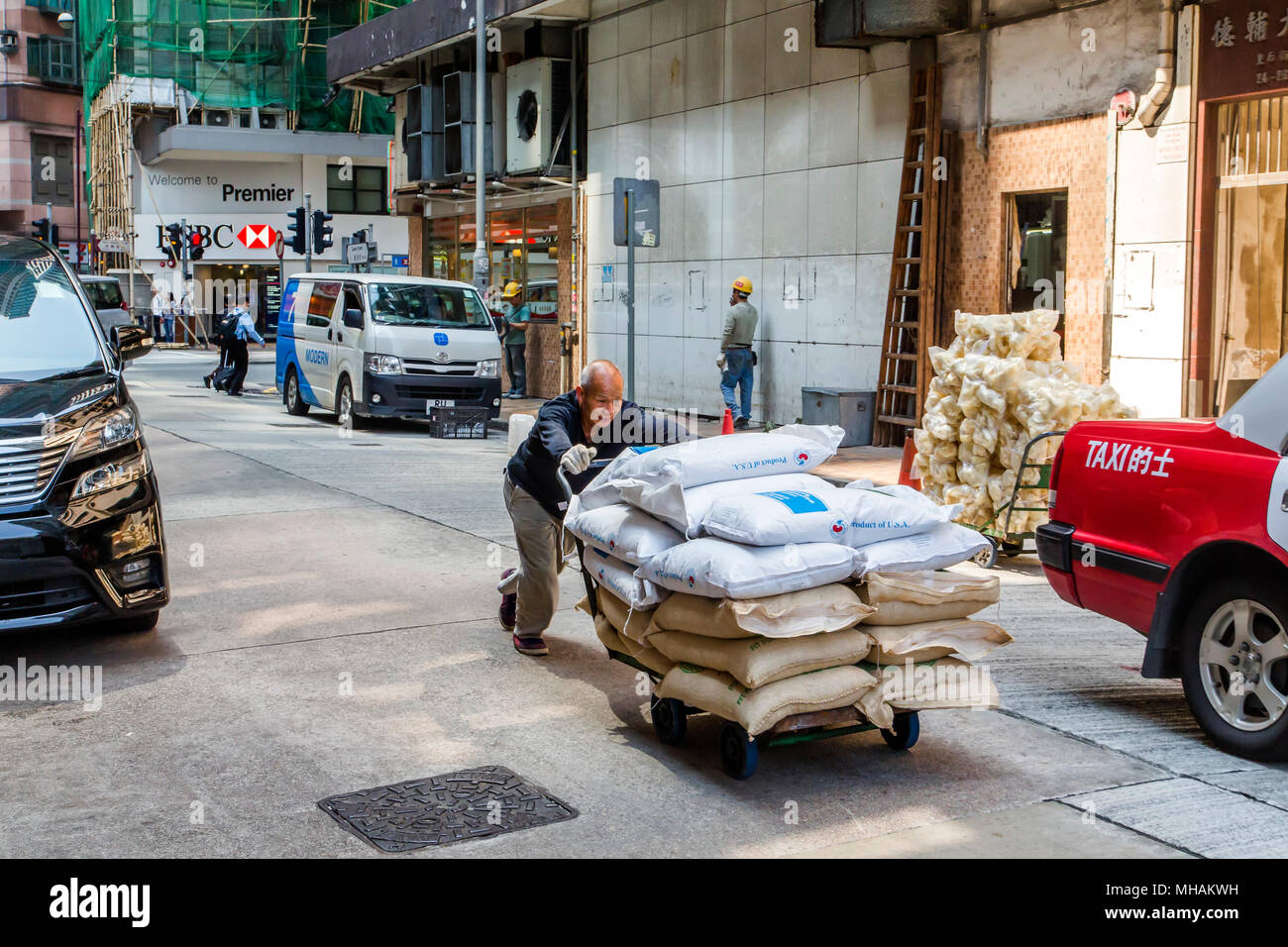 Un anziano uomo asiatico spinge un carrello caricato con sacchi di merci attraverso una strada di Hong Kong Island. Alcuni sacchi sono contrassegnati come prodotto di U.S.A. Foto Stock