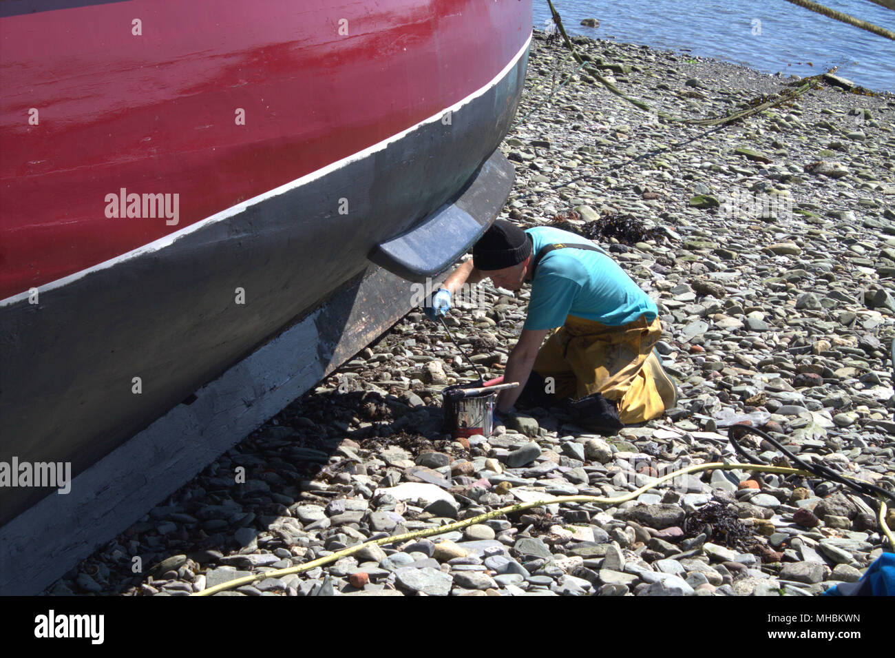 Fisherman pittura antivegetativa sulla carena della sua barca da pesca, approfittando della bassa marea e caldo secco meteo. Foto Stock