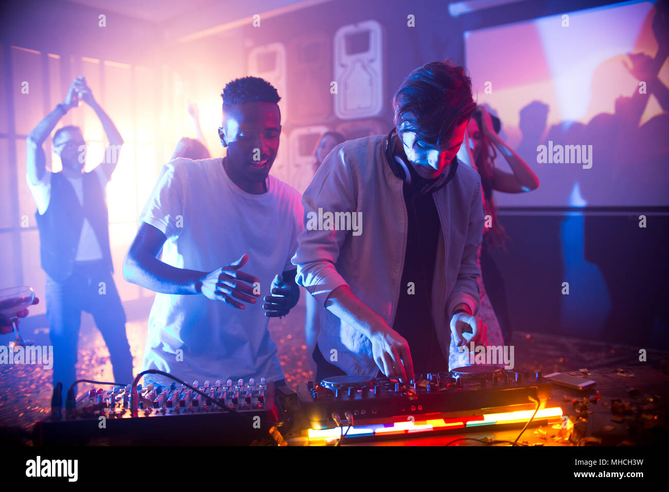 Vita ritratto di bel giovane DJ permanente al mixer rendendo la musica durante la festa notturna in club, scena illuminata da luci da palco con persone che ballano in ba Foto Stock