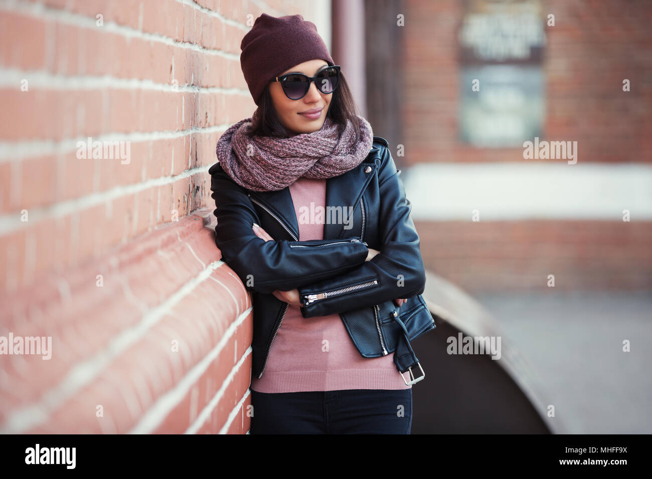 Moda giovane donna che indossa gli occhiali da sole e sciarpa beanie appoggiato su di un muro di mattoni elegante modello femminile in pelle nera camicia outdoor Foto Stock