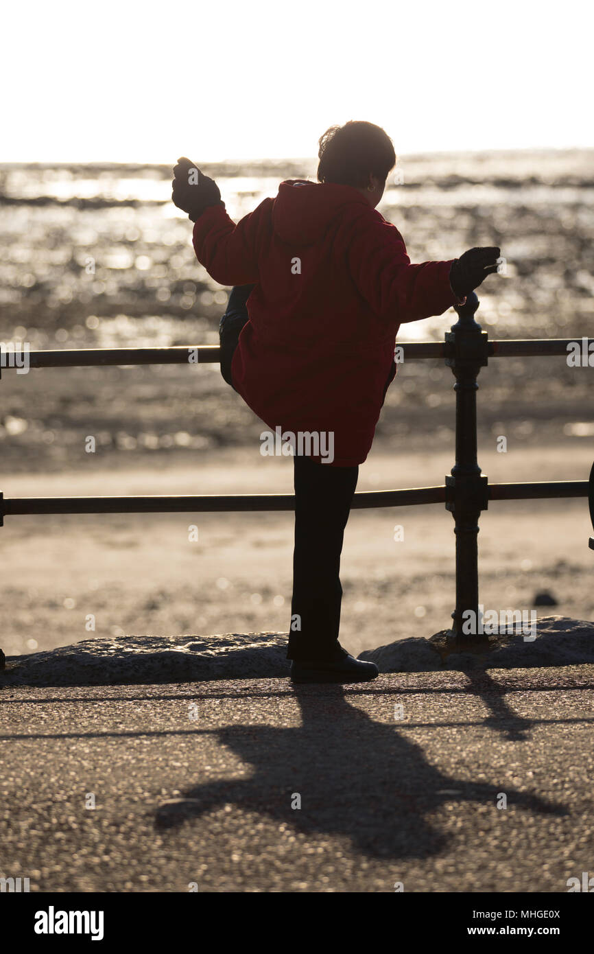 Un anziano le donne cinesi facendo la mattina presto il tai chi esercizio utilizzando le ringhiere su teh lungomare per supporto a Llandudno, Galles Foto Stock