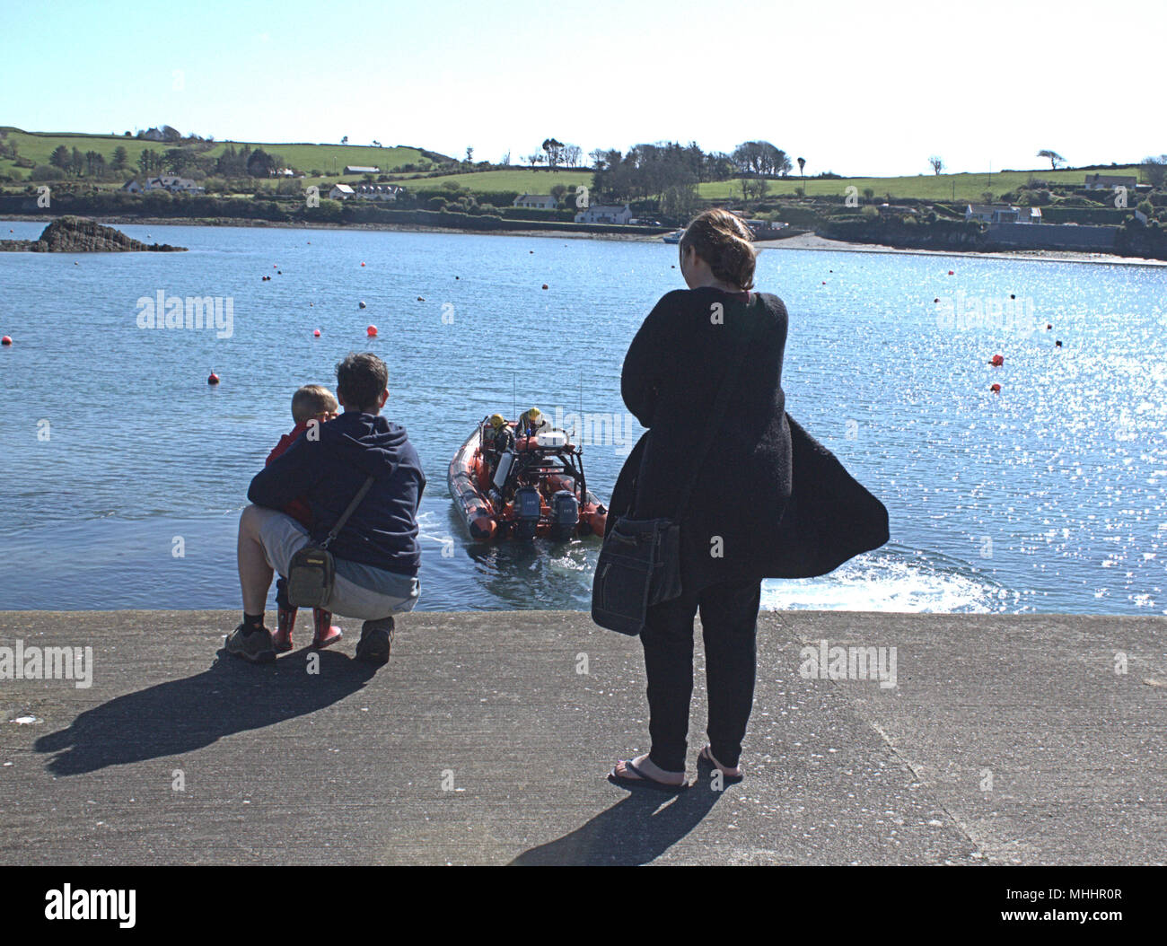 Famiglia coppia in vacanza con un piccolo ragazzo guardando la guardia costiera irlandese la barca di salvataggio di essere lanciato dalla fine di un pontile in Irlanda. Foto Stock