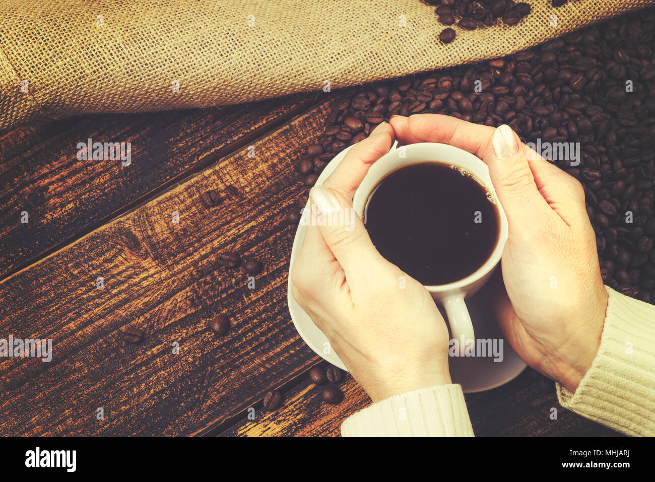 Concetto di degustazione del caffè in stile rustico - la donna detiene una tazza di caffè nelle sue mani su un tavolo di legno con fagioli fuoriuscito da un sacco di tela (vi Foto Stock