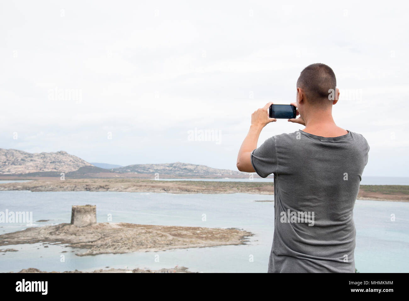 Primo piano di un giovane uomo caucasico visto da dietro di scattare una foto con il suo smartphone, della Torre della torre della Pelosa e il mar mediterraneo in Foto Stock