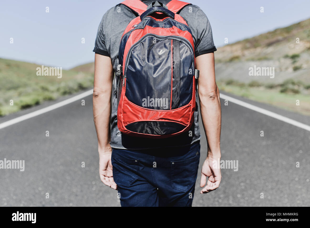 Primo piano di un giovane uomo caucasico, visto da dietro, portando uno zaino camminando lungo una strada secondaria Foto Stock