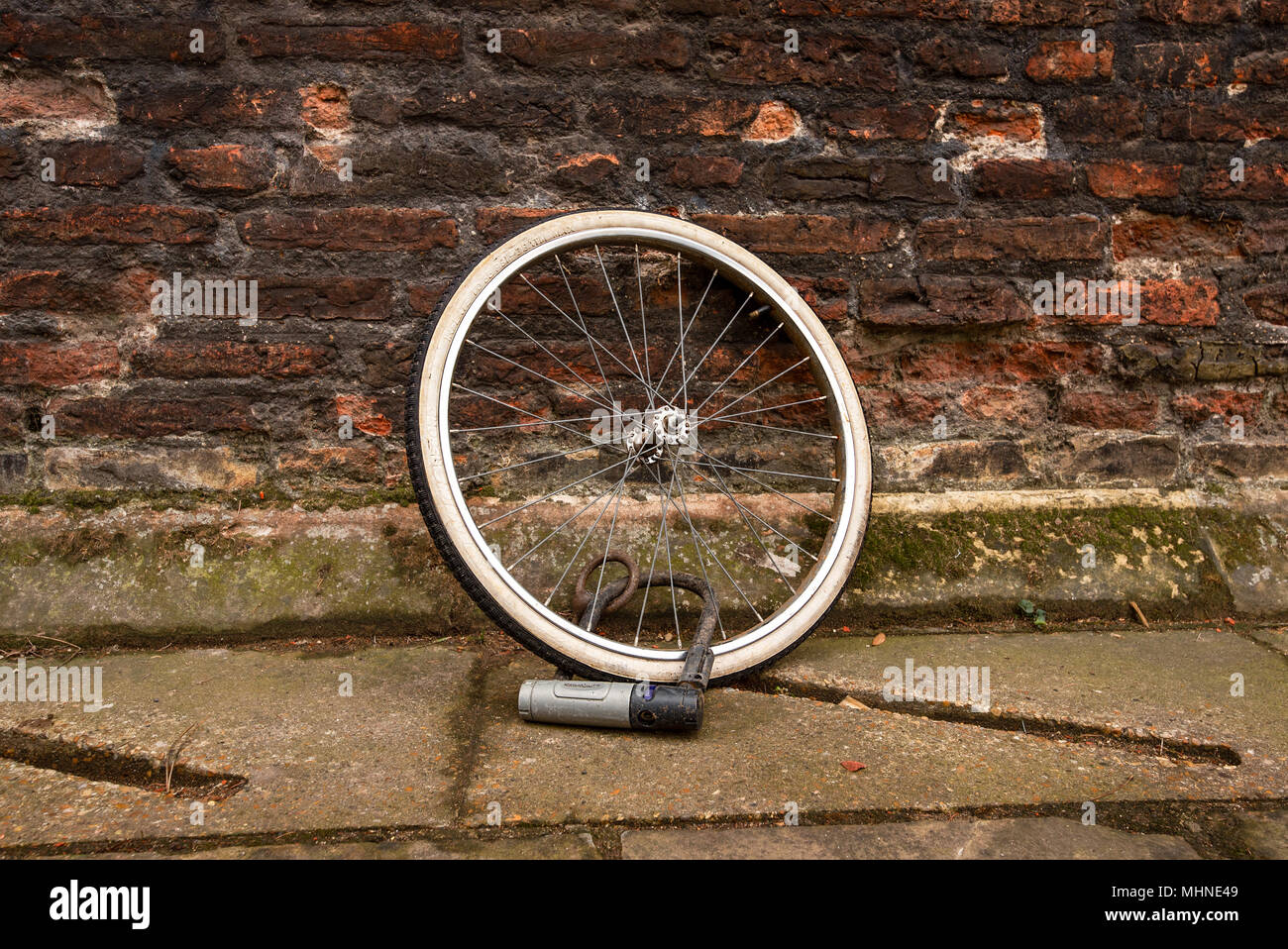 Una solitaria la ruota di bicicletta con pneumatici ma manca il suo moto è securly bloccato con D lock contro un vecchio rosso un muro di mattoni. Foto Stock